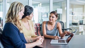 three people siting at a desk with a laptop talking