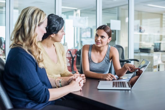 three people siting at a desk with a laptop talking